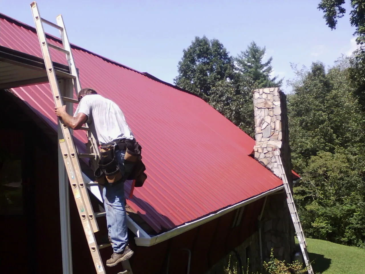 Roofer on a ladder beside a red metal roof and stone chimney — Soffit & Fascia Repair by Roofing Brothers in Willimantic